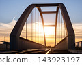 Close view of arched steel railway bridge in Magdeburg, Germany, silhouetted against glowing sunset sky. Vertical suspension rods and dark metal girders frame warm evening light above empty tracks 143923197
