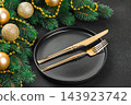 Elegant Christmas table setting with gold cutlery on black plate and decorated spruce branches 143923742