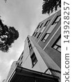 Monochrome geometric building facade between silhouetted trees under cloudy sky 143928257