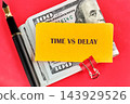 A yellow note reading Time vs Delay rests on US dollar bills beside a black pen on a red surface 143929526