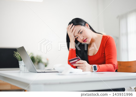 Woman in red shirt worried while checking credit card bill at table with laptop and receipt 143931996