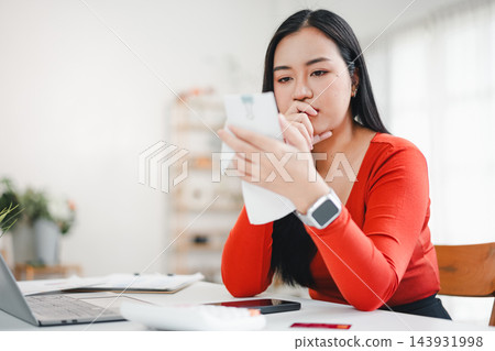 Young woman in red sweater reviewing receipt at home desk, thoughtful expression. Young woman in red sweater reviewing receipt at home desk, thoughtful expression. 143931998