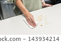 Woman cleaning crumbs from kitchen table after slicing cake, homemade baking process, food preparation routine, cozy home cooking lifestyle. 143934214