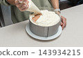 Woman spreading white cream over sponge cake in metal ring on kitchen table, homemade baking process, dessert decoration stage, cozy home cooking lifestyle. 143934215