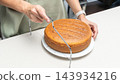 Woman cutting piece of sponge cake with knife on white plate in kitchen, homemade baking process, food preparation routine, cozy home cooking lifestyle. 143934216