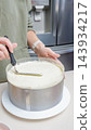 Person in green sweater smooths frosting on a round cake using a spatula in a modern kitchen with stainless steel appliances visible in the background 143934217