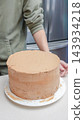Person decorating a round chocolate cake with smooth frosting on a white cake stand in a modern kitchen setting 143934218