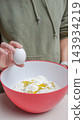 Person holding a white egg above a bowl filled with flour and yolk, wearing a green hoodie in a kitchen setting 143934219