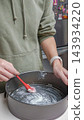Person in green hoodie applies butter to a round baking pan with a red spatula in a modern kitchen setting with stainless steel appliances 143934220
