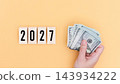 Hand holding a stack of United States dollar bills beside wooden number blocks displaying 2027 year on a warm orange background, financial concept 143934222
