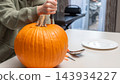 Person carving a large orange pumpkin with a knife on a kitchen table, plate and utensils visible in a well-lit indoor setting 143934227