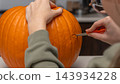 Person carving a bright orange pumpkin for Halloween with a small knife, seated at a table in a cozy indoor setting with kitchen elements visible 143934228