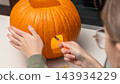 Person carving a Halloween pumpkin with a small knife, showing intricate details and orange flesh on a light-colored countertop in a modern kitchen setting 143934229