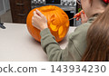 Child carving a pumpkin for Halloween with a small knife on a kitchen countertop, kitchen appliances visible in the background 143934230