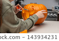 Person carving a pumpkin for Halloween in a kitchen, with a focus on the intricate design being created and kitchen appliances visible in the background 143934231