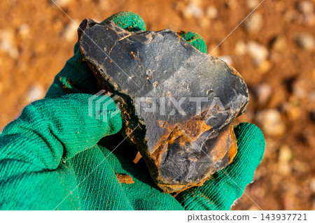 Gloved hand holding a raw piece of dark flint stone found in an industrial quarry, featuring unique mineral textures and geological patterns against a blurred earthy background Gloved hand holding a raw piece of dark flint stone found in an industrial quarry, featuring unique mineral textures and geological patterns against a blurred earthy background 143937721