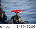 Red Metal Valve Wheel Mounted on Industrial Piping Against Blurred Water Background 143943286