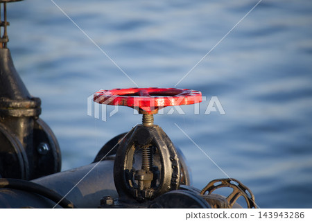 Red Metal Valve Wheel Mounted on Industrial Piping Against Blurred Water Background 143943286