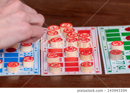 Player's hand marks a matching number on a bingo card with a wooden token during a casual indoor game, close up showing multiple cards and the thrill of winning Player's hand marks a matching number on a bingo card with a wooden token during a casual indoor game, close up showing multiple cards and the thrill of winning 143943394