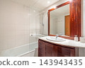 Modern bathroom interior with wooden vanity, large mirror, glass shower, white bathtub, chrome fixtures, bright recessed lighting, and minimalist contemporary design 143943435