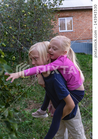 Two blond children, a boy giving a piggyback ride to a girl, are reaching for green apples hanging from a tree in a lush green garden with a brick house in the background Two blond children, a boy giving a piggyback ride to a girl, are reaching for green apples hanging from a tree in a lush green garden with a brick house in the background 143943530