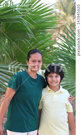 Mother and son smile together outdoors, standing close beneath arching palm fronds. Relaxed family portrait reflects loving maternal bond, with both facing camera in bright daylight. Mother and son smile together outdoors, standing close beneath arching palm fronds. Relaxed family portrait reflects loving maternal bond, with both facing camera in bright daylight. 143945020