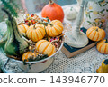 striped orange pumpkins and green gourd in white colander on lace table, cozy autumn harvest decoration featuring seasonal squash near vintage candlestick 143946770