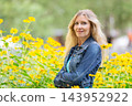 Smiling senior woman in denim jacket surrounded by bright yellow flowers under soft natural light 143952922