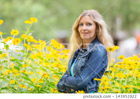 Smiling senior woman in denim jacket surrounded by bright yellow flowers under soft natural light 143952922