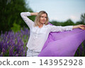 Woman in a flowing white shirt poses gracefully with a purple fabric in a field of blooming purple flowers on a sunny day 143952928