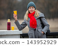 Woman enjoys warm drink in winter park while wearing a grey coat and red scarf 143952929
