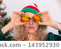 An elderly woman in a Christmas hat with a playful expression holds oranges above her eyes during Christmas 143952930