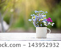Flowers in white cup sit on table in garden during daytime with sunlight shining through plants in background 143952932