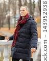 Elderly man enjoying a cool autumn day in a park while wearing a warm coat and scarf with a cane 143952938