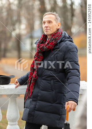Elderly man enjoying a cool autumn day in a park while wearing a warm coat and scarf with a cane Elderly man enjoying a cool autumn day in a park while wearing a warm coat and scarf with a cane 143952938