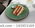 Slice of layer carrot cake with cream frosting served on green plate with dessert spoon on white table outdoor 143953169