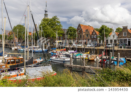 Horizontal perspective of town hall clock tower harbor marina sailboats traditional Dutch brick houses wooden posts blue sky Veere Zeeland Netherlands European travel commercial 143954430