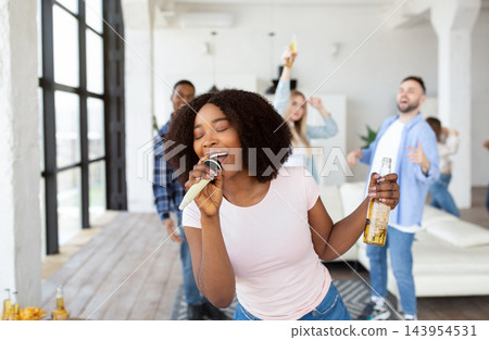 Friendship, domestic entertainments. Happy black woman with bottle of beer and microphone performing song, singing karaoke on party with her diverse friends, having fun at home Friendship, domestic entertainments. Happy black woman with bottle of beer and microphone performing song, singing karaoke on party with her diverse friends, having fun at home 143954531