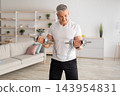 Workout during lockdown. Athletic middle-aged man doing dumbbell workout at home, looking at his biceps, copy space. Mature man training in living room interior 143954831