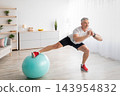 Athletic mature man doing side lunges, using fitness ball, working out at home in living room interior, empty space. Man exercising, training in the morning during quarantine 143954832