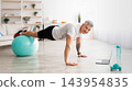 Domestic online training. Athletic mature man standing in plank with legs on fitness ball, working out and watching sports tutorial videos on laptop, panorama 143954835