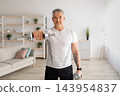 Domestic training with weights. Positive mature man doing exercises with dumbbells, strengthening his body at home. Senior sportsman working on his biceps muscles, staying healthy 143954837