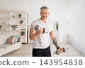 Sporty mature man training with dumbbells at home, working on arms strength, looking and smiling at camera, copy space. Sportsman lifting dumbells up over living room interior 143954838
