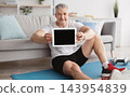 Happy mature man sitting on fitness mat at home and showing digital tablet with blank screen, demonstrating place for your ad or website design, mockup, empty space 143954839