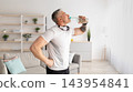 Stay hydrated. Mature man drinking water during home workout, standing in living room with headphones on neck, panorama. Home training, fitness hydration, sporty lifestyle concept 143954841
