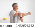Home training. Sporty mature man stretching hands, warming up before exercising in living room interior at home and looking aside at window, copy space 143954842