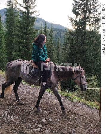 A gray horse carries a woman past tall evergreen trees, showing the steady pace of a recreational ride through a mountain landscape 143955028