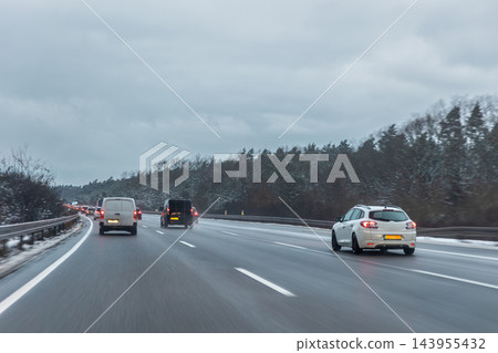Winter traffic moves slowly along German highway under heavy gray clouds, with cars and van forming long queue. Wet asphalt, snowy shoulders and blurred motion convey cold seasonal commute 143955432