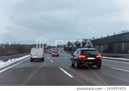 Winter traffic moves slowly along German highway under heavy gray clouds, with cars and van forming long queue. Wet asphalt, snowy shoulders and blurred motion convey cold seasonal commute Winter traffic moves slowly along German highway under heavy gray clouds, with cars and van forming long queue. Wet asphalt, snowy shoulders and blurred motion convey cold seasonal commute 143955433