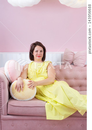Young woman reclining on pink sofa in pastel living room wearing yellow dress and purple sandals, eyes closed in calm fashion pose Young woman reclining on pink sofa in pastel living room wearing yellow dress and purple sandals, eyes closed in calm fashion pose 143956688